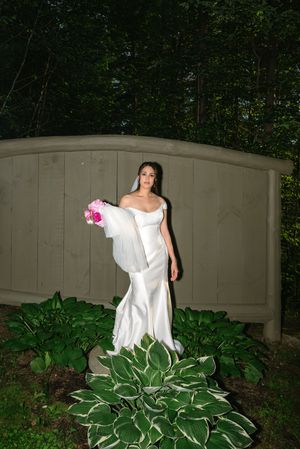 Bride in white off-shoulder mermaid gown holding pink bouquet, standing by wooden arch surrounded by lush green hosta plants.