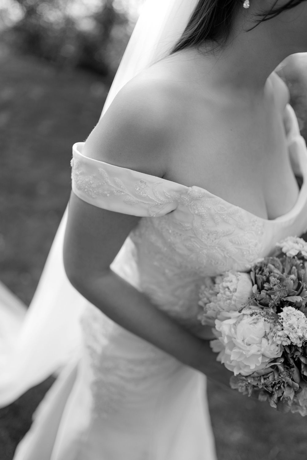 Black and white photo of a bride in an off-shoulder lace wedding gown holding a bouquet of flowers.
