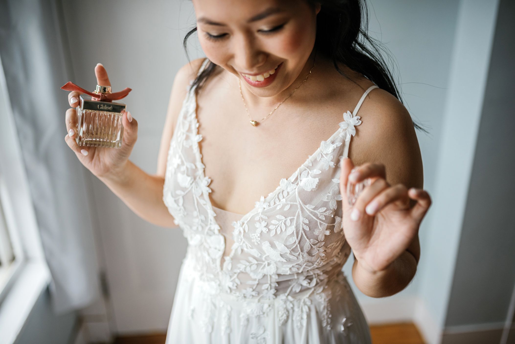 Person in white lace wedding dress holding a perfume bottle near a window.