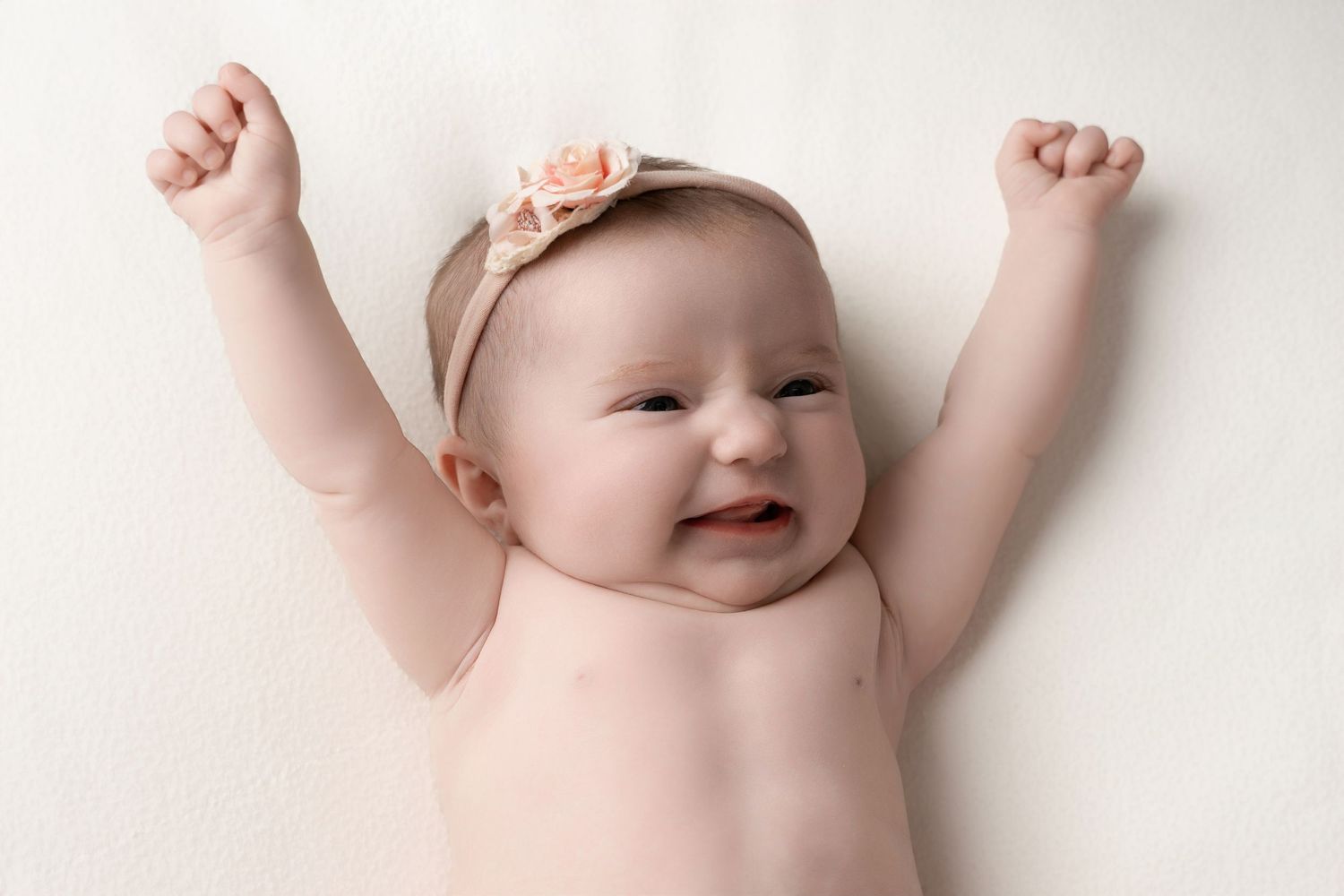 Happy baby wearing pink headband raises arms up joyfully while lying on white background.