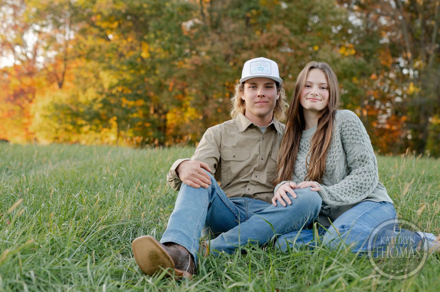 A couple relaxes together on grass with fall foliage in the background.