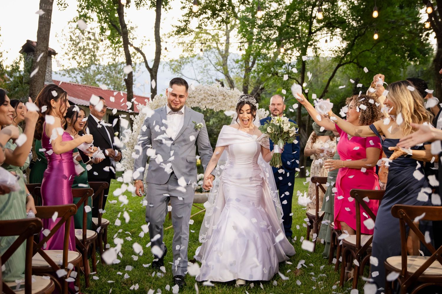 Wedding guests toss flower petals as newlyweds walk down garden aisle.