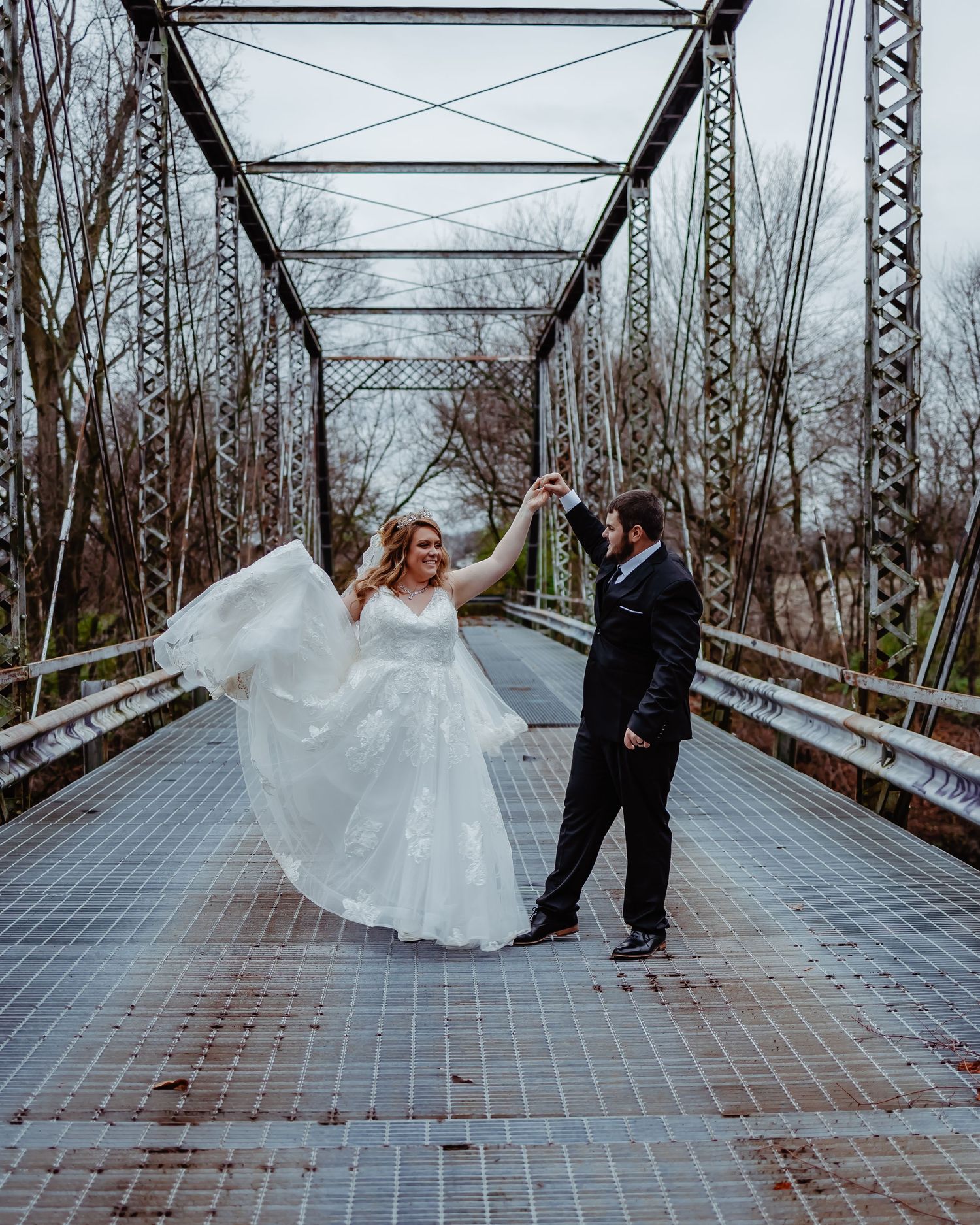 A couple celebrates their wedding day with a twirling dance pose on a rustic metal bridge surrounded by bare winter trees.