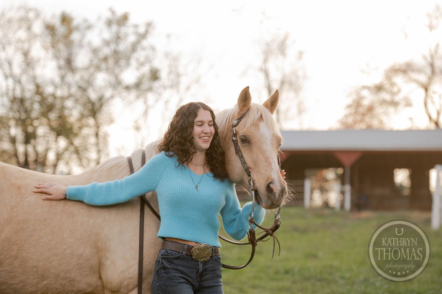 Person in blue sweater poses with palomino horse in autumn pasture.