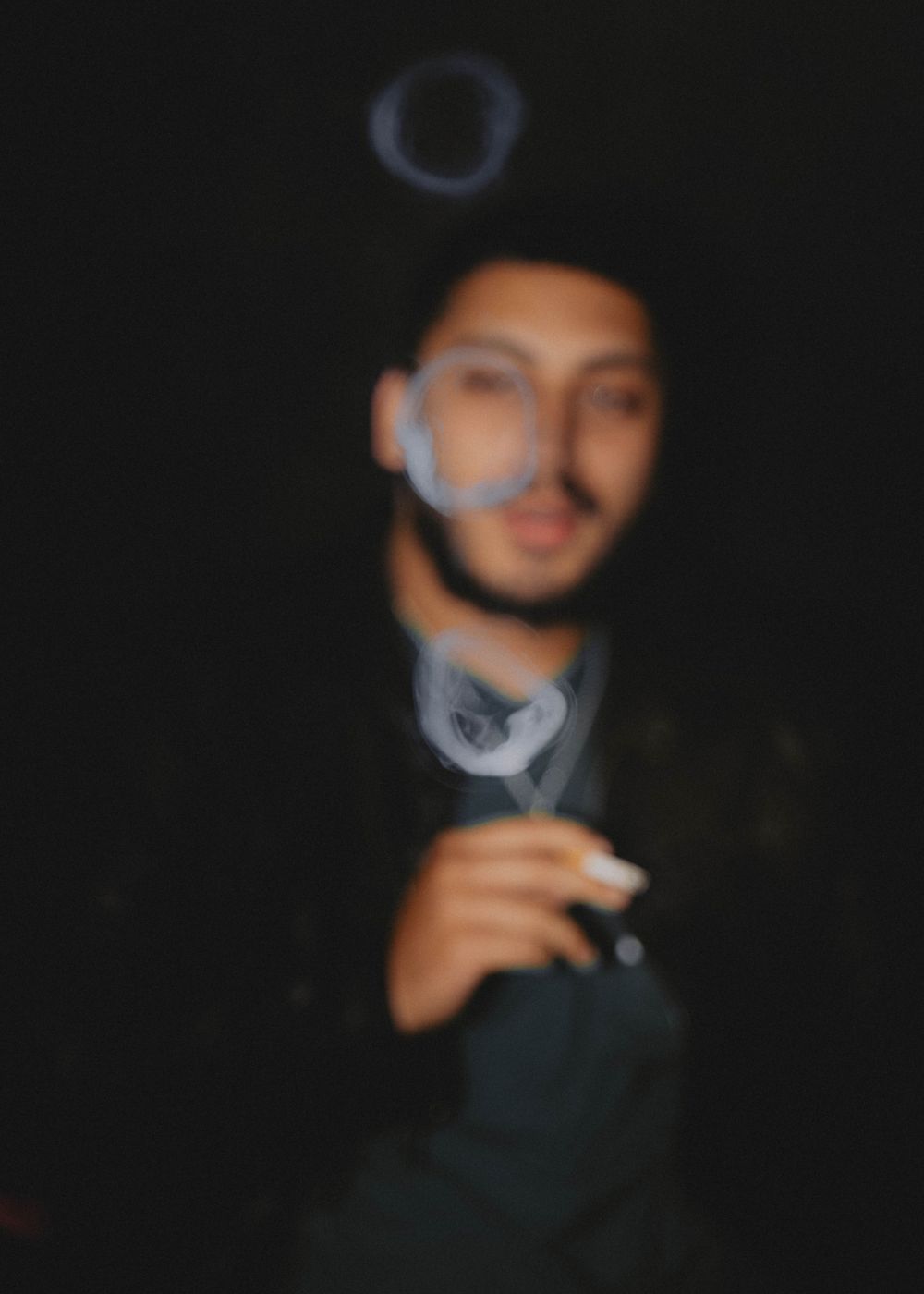 A blurry nighttime portrait showing someone wearing a baseball cap and holding a cigarette in dark lighting.