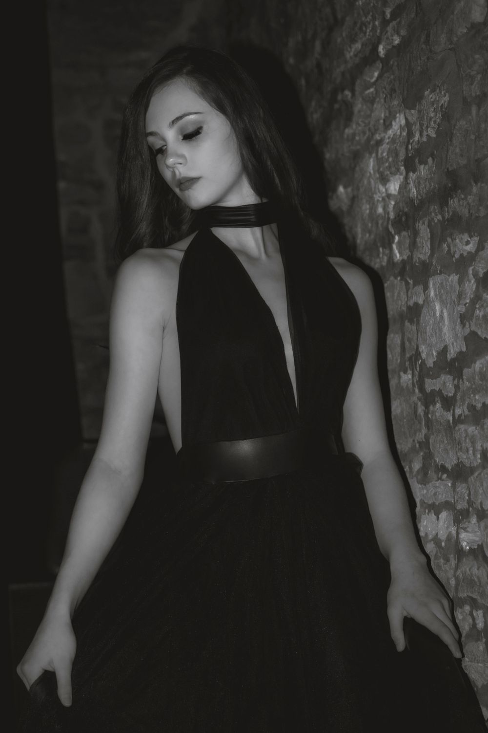 Artistic black and white portrait of someone in an elegant black dress with choker necklace against stone wall.