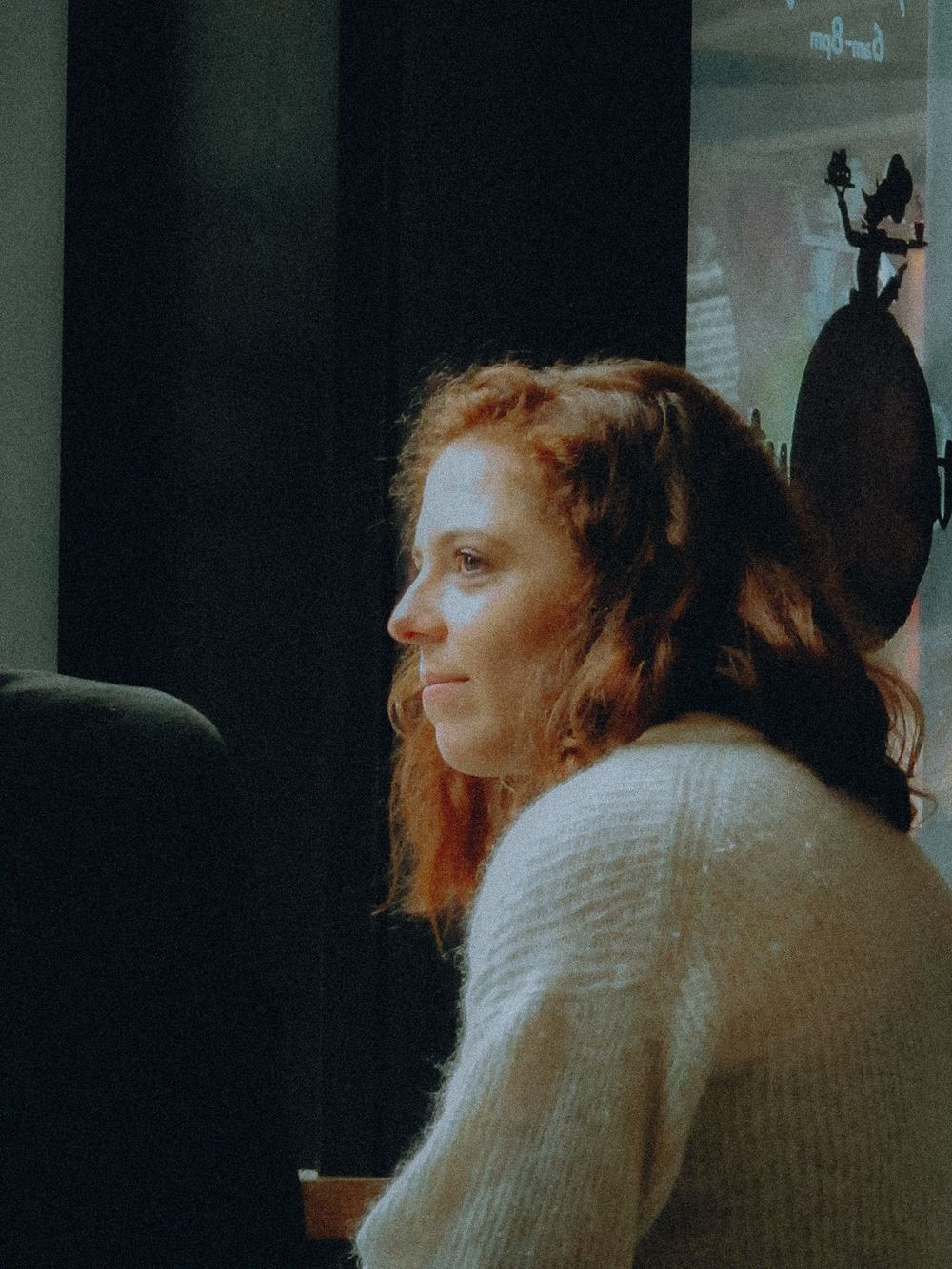 Person with long auburn hair in a cream sweater sits in profile view by a window in soft lighting.