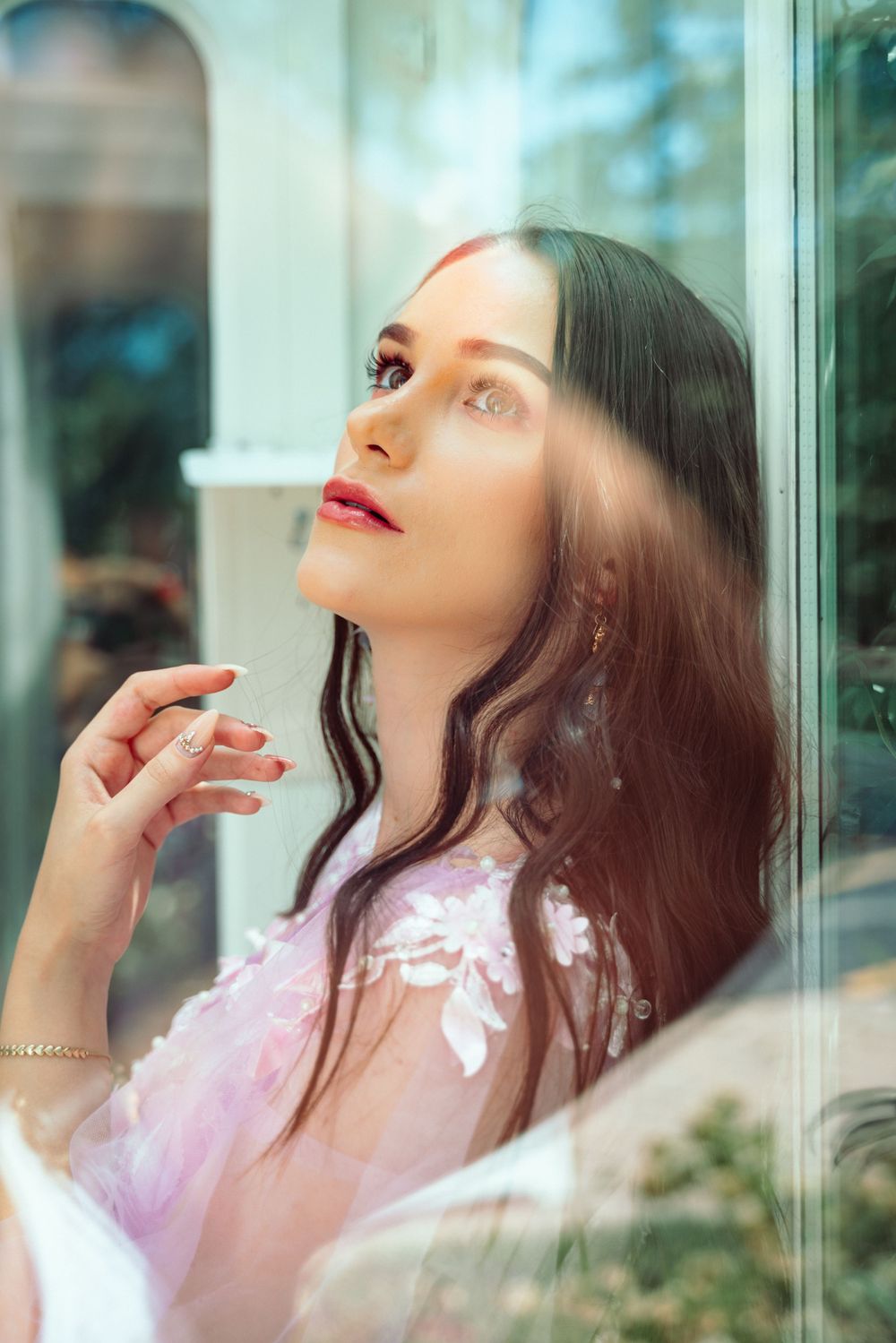 Ethereal portrait through window glass showing soft pink clothing and dreamy lighting with reflections.