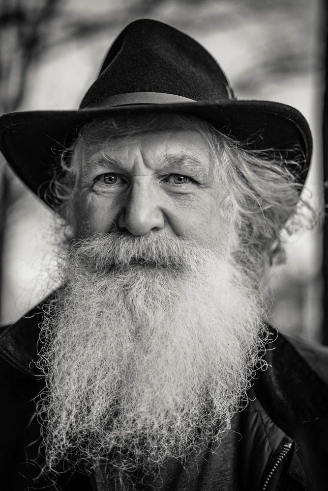 Black and white portrait of elderly man with long white beard wearing dark hat.