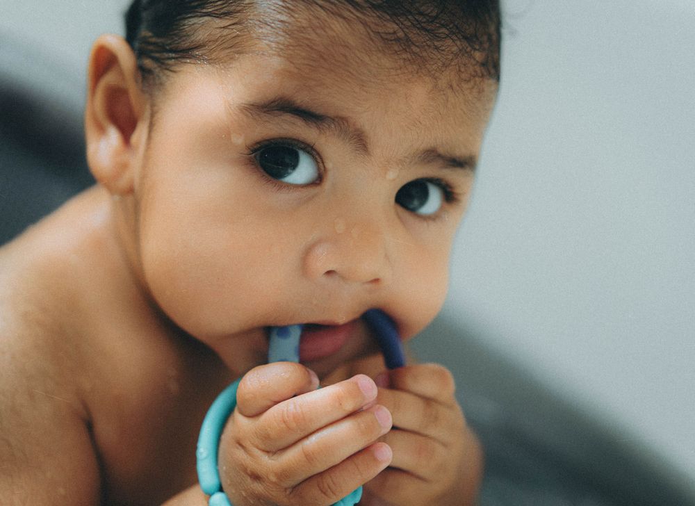 Close up of a young child biting on a light blue silicone teething toy against a plain background.
