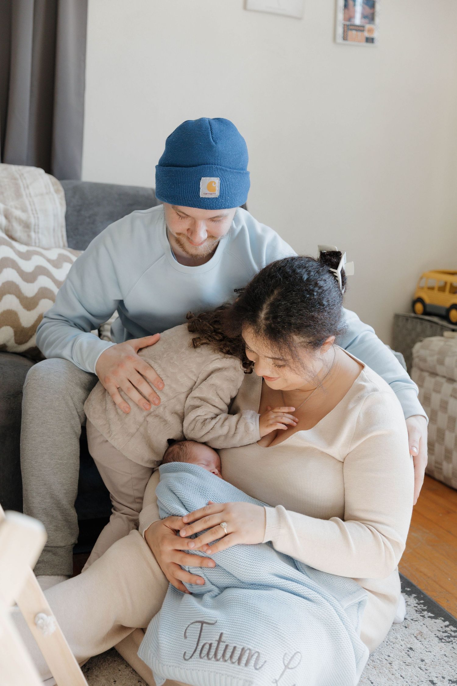 A couple lovingly embraces their newborn baby while sitting together on a couch at home.
