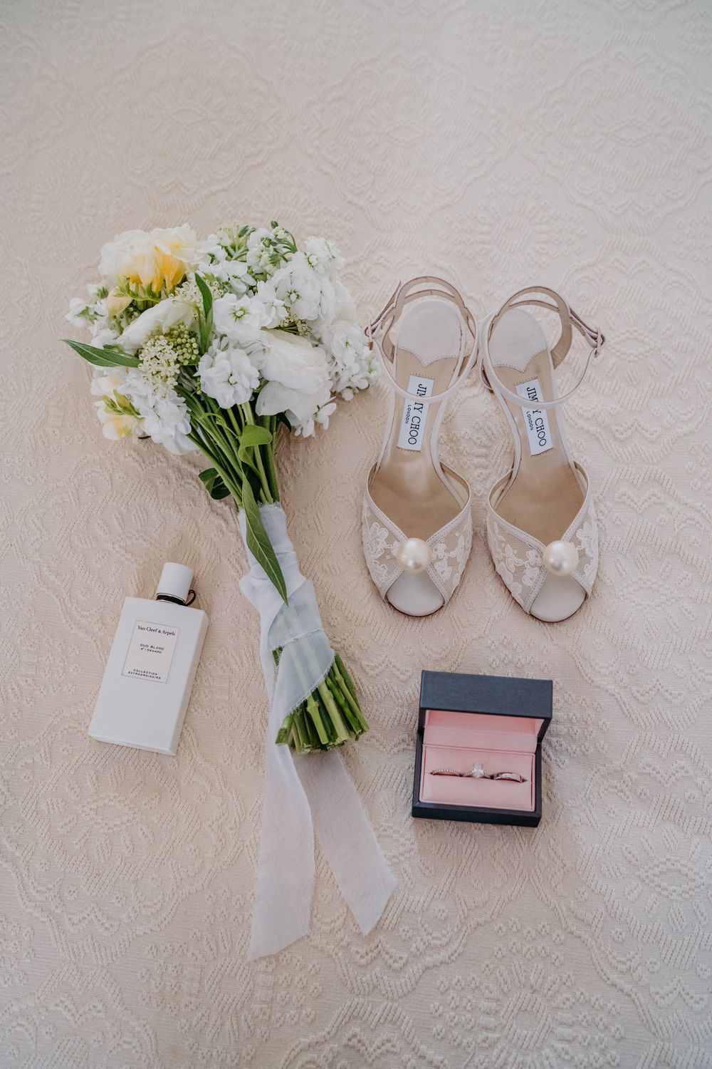 Wedding day details including white floral bouquet, Jimmy Choo sandals, perfume bottle and ring box arranged on lace.