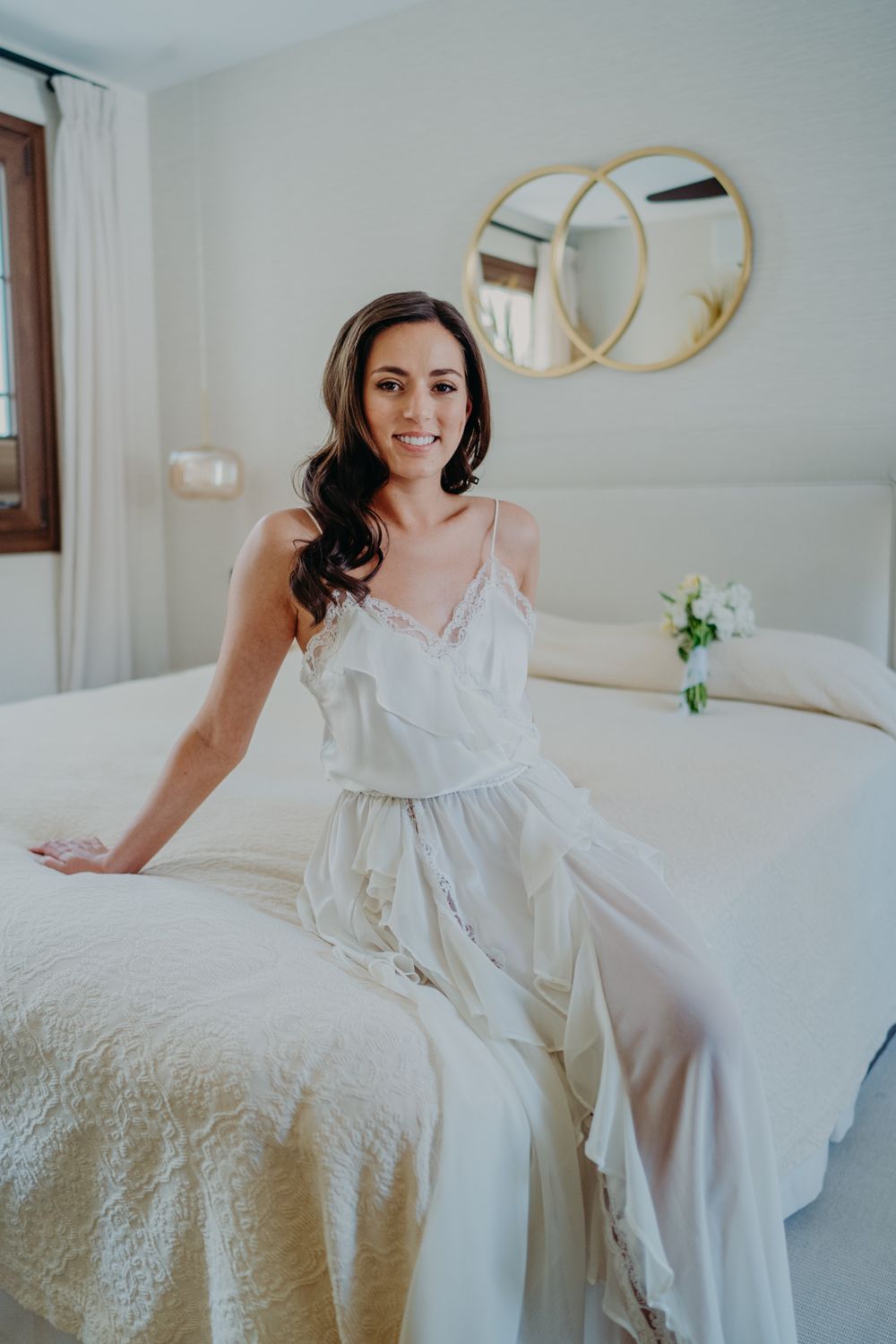 Woman in white satin dress sits on bed with circular gold mirrors on wall in bright bedroom setting.