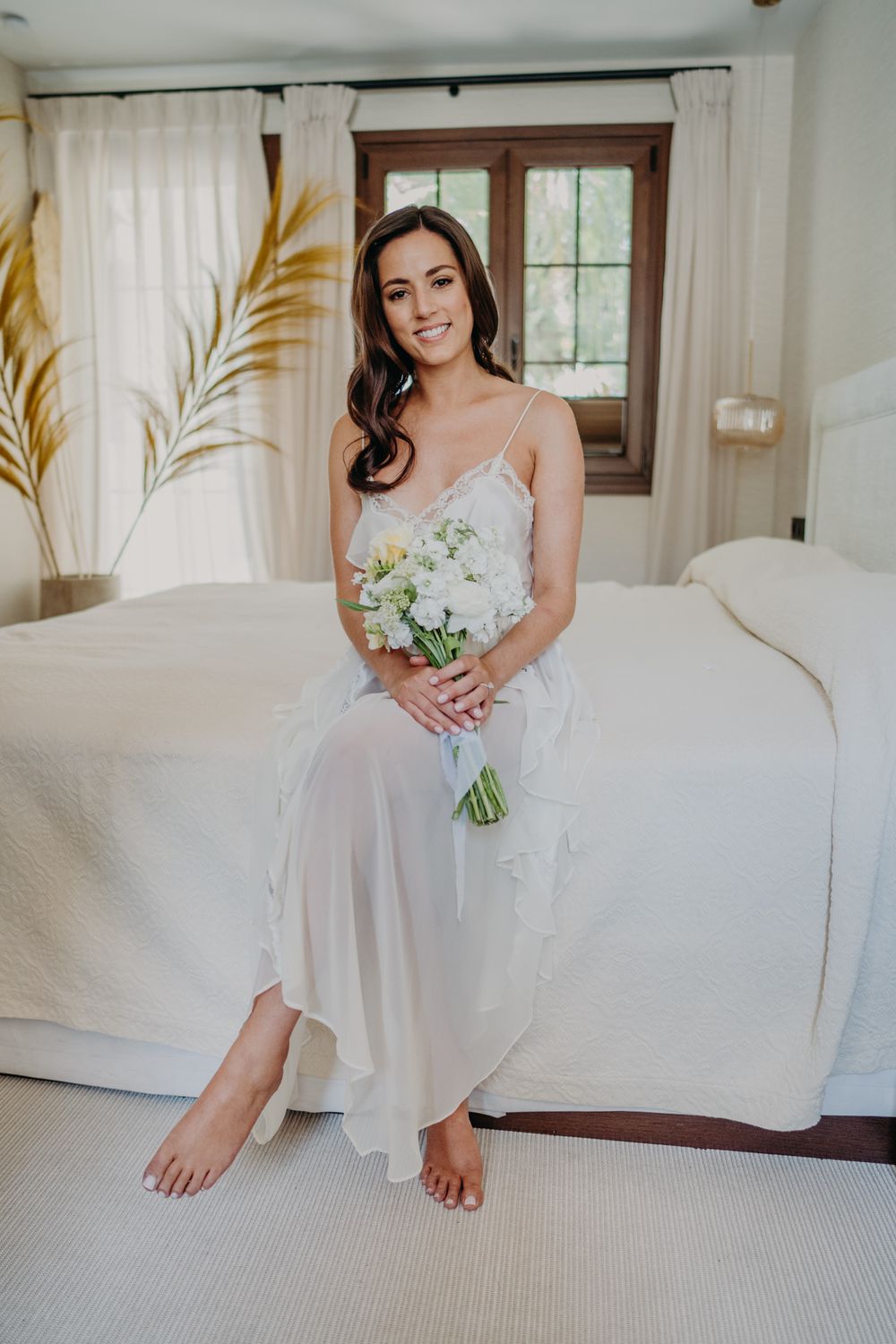 A bride wearing a white satin gown sits barefoot on a bed holding a bouquet of white flowers.
