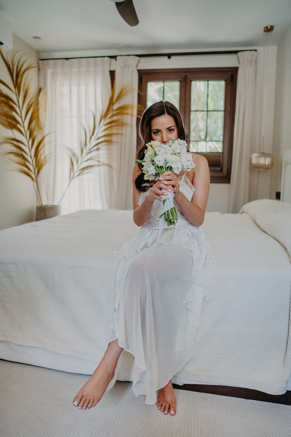 A bride in a flowing white dress sits barefoot on a bed holding a bouquet of white flowers near a window.