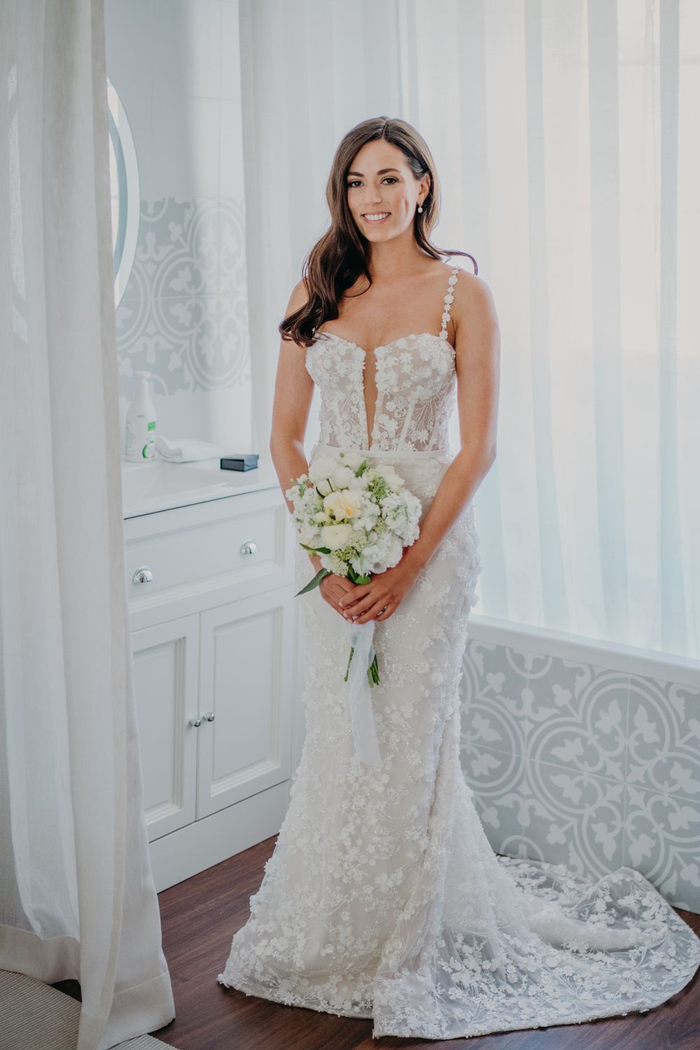 A bride in a fitted lace wedding gown holds a white rose bouquet while posing in an elegant dressing room.