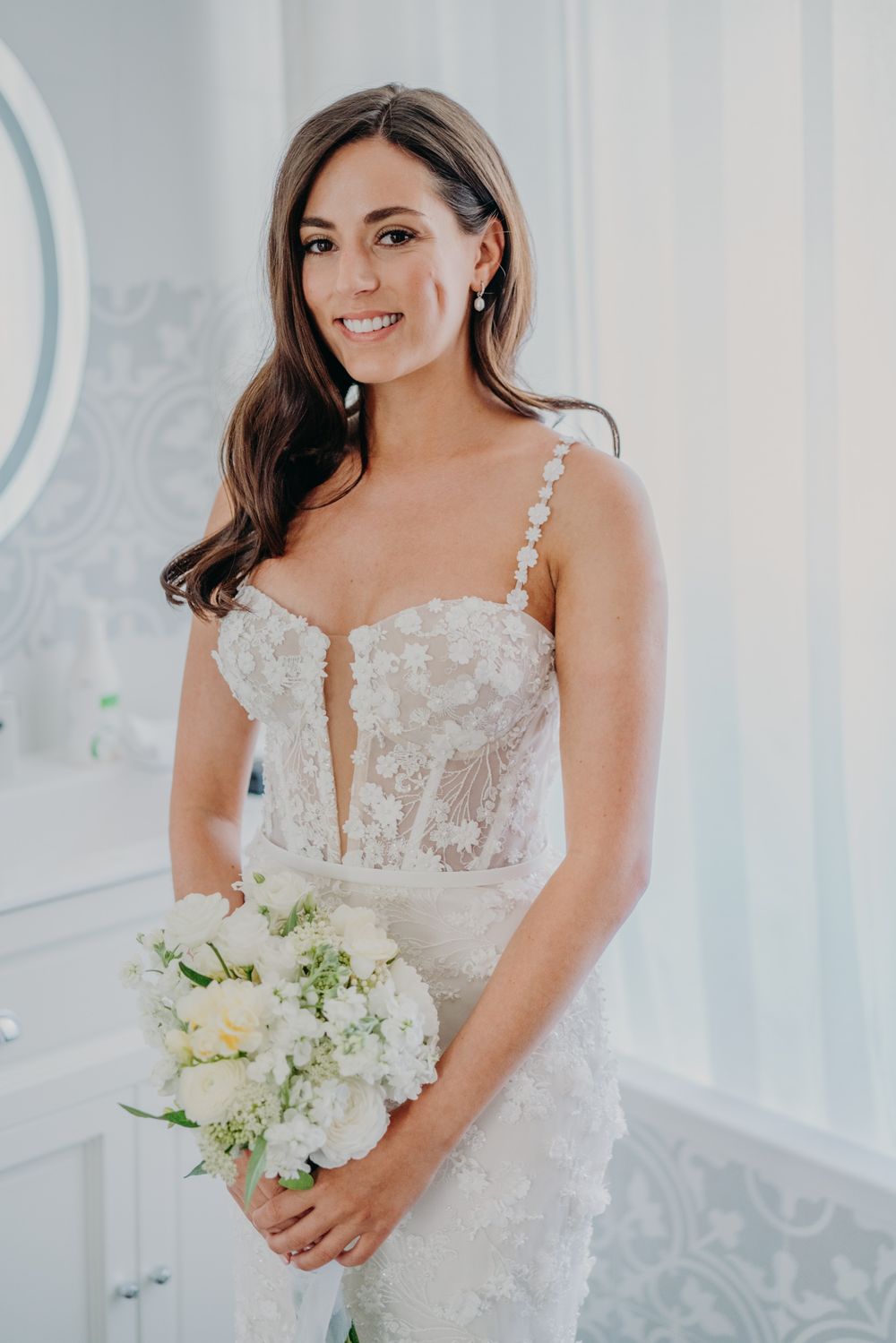 Beautiful bride in lace corset wedding dress holds white and ivory floral bouquet in bright room.
