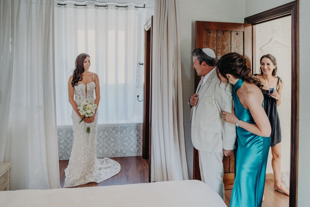 In a sunlit room with white curtains, people stand in elegant formal attire preparing for a special occasion.