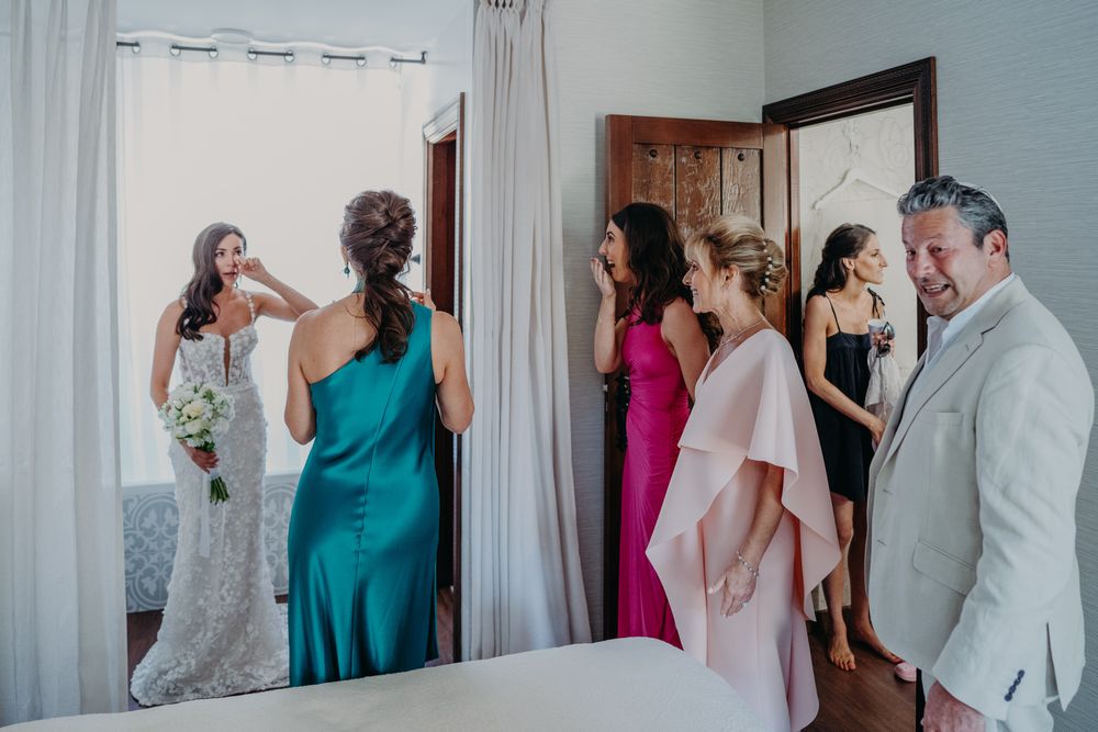 Wedding party members in colorful dresses gather around a mirror in a getting-ready room on the big day.