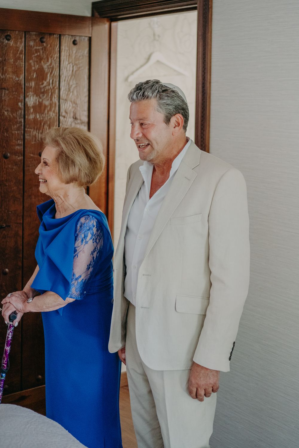 Two people share a happy moment wearing formal attire, a royal blue dress and light colored suit in an indoor setting.