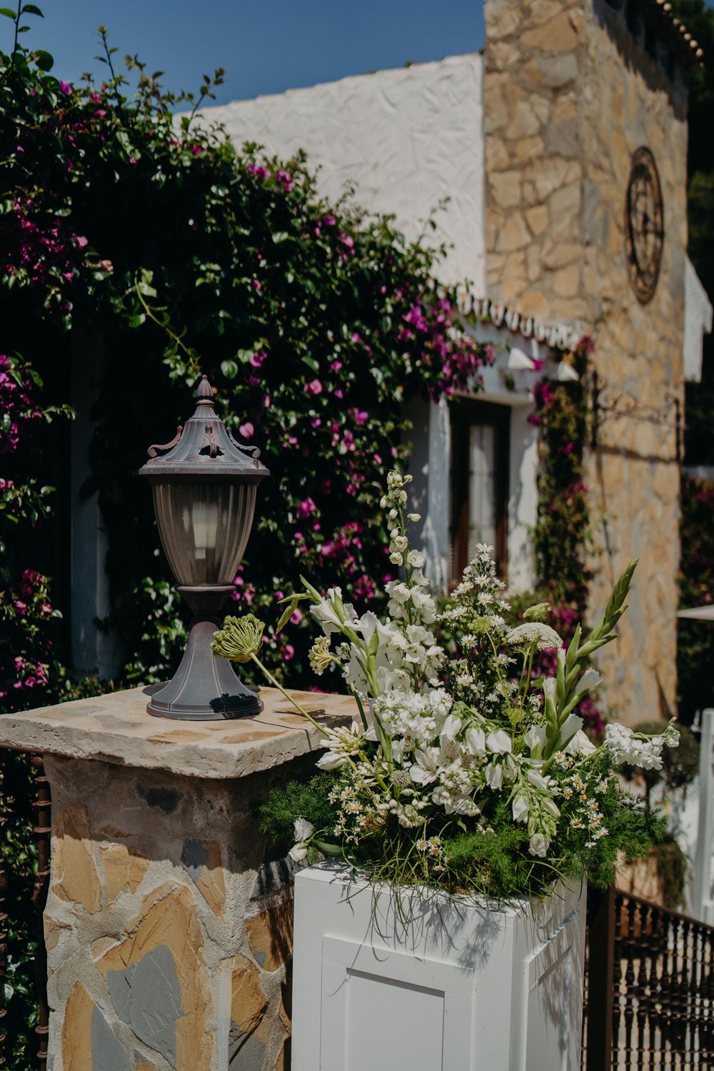 Beautiful stone wall entrance decorated with pink bougainvillea flowers and an elegant lamp post with white floral arrangements.