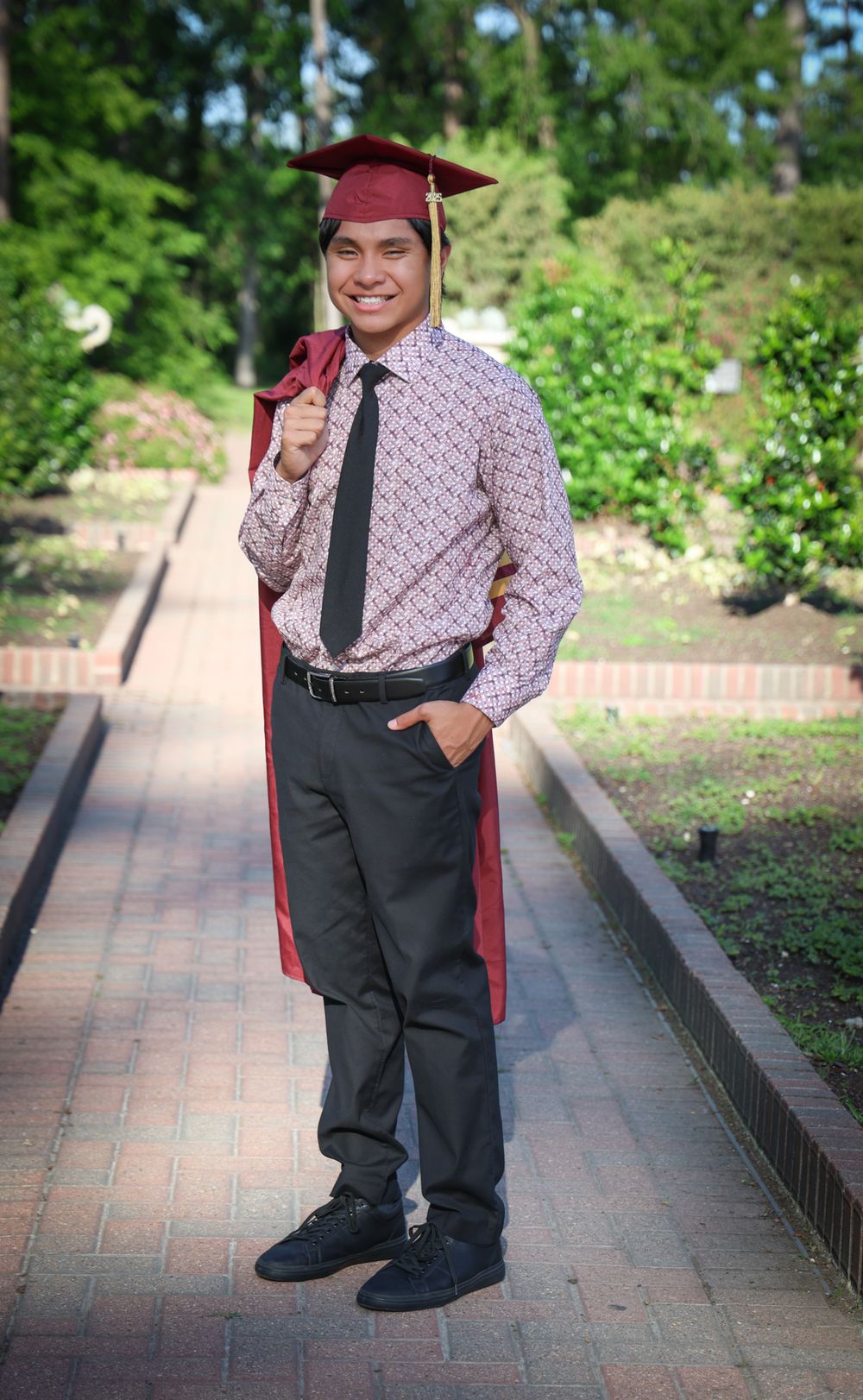 Graduate in burgundy cap and gown poses on brick pathway surrounded by greenery.