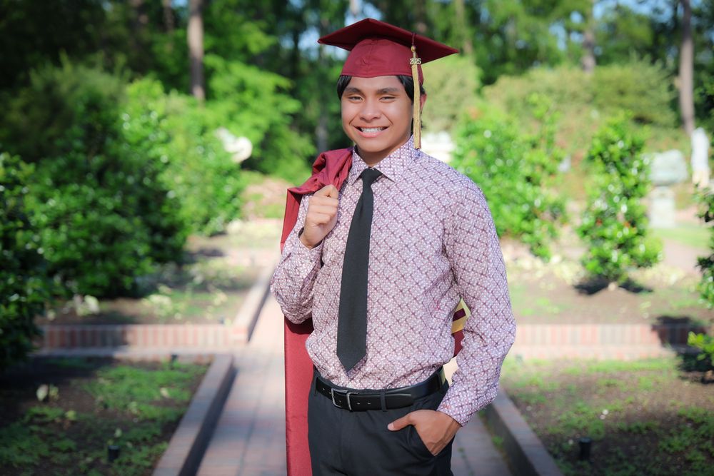 Graduate in burgundy cap and printed shirt poses outdoors on brick walkway.