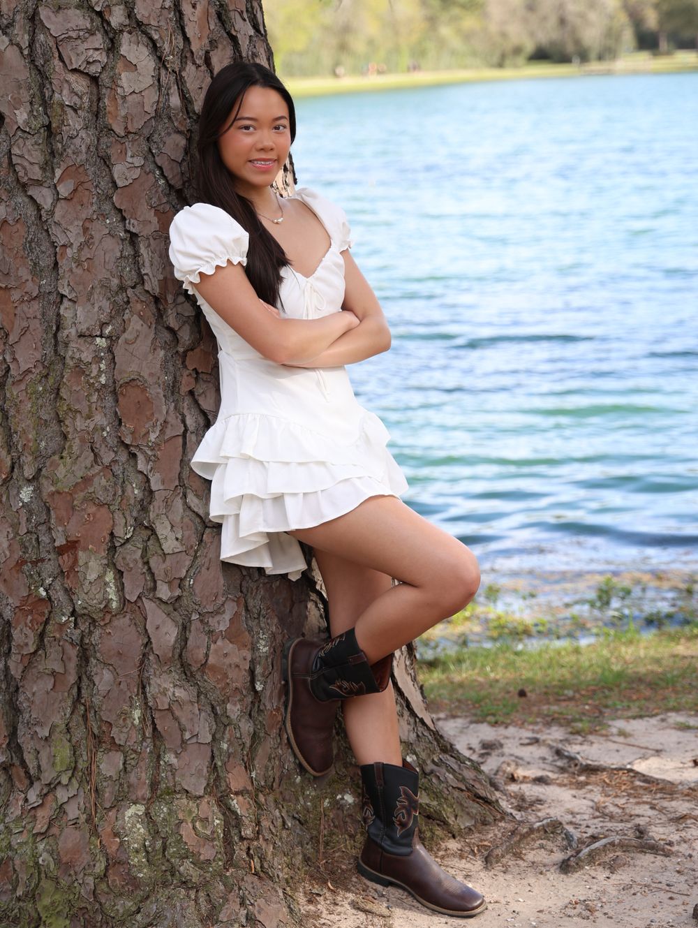 Outdoor photoshoot by a lake featuring white dress and boots against a large tree trunk.