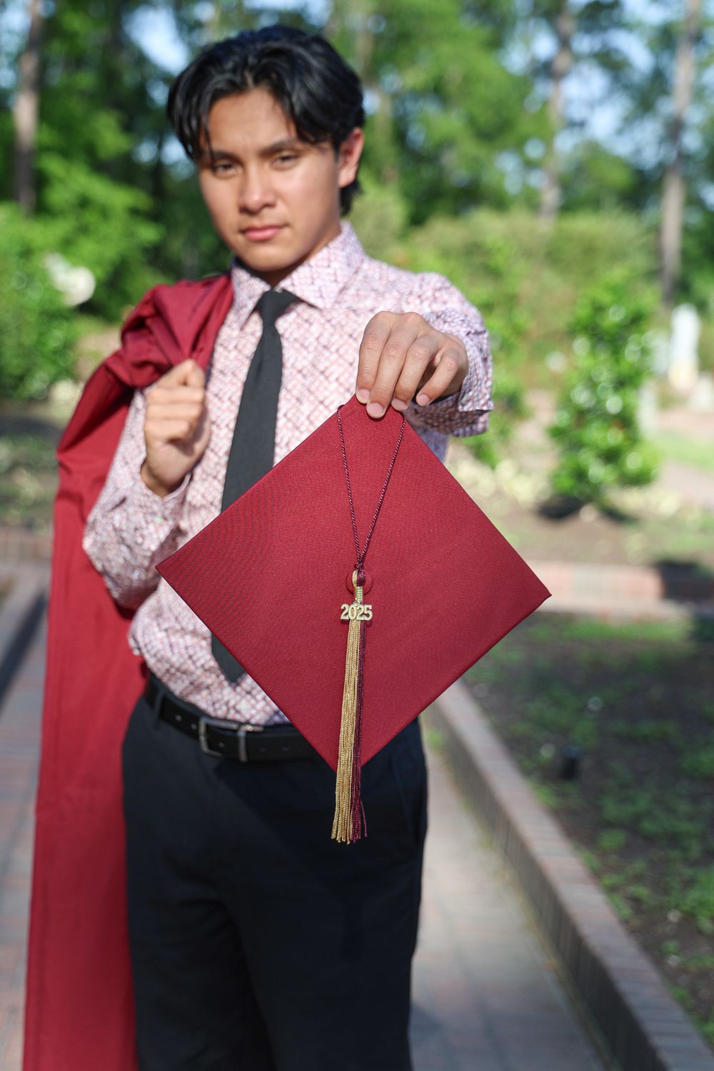 Graduate displays burgundy graduation cap with tassel against brick pathway backdrop.