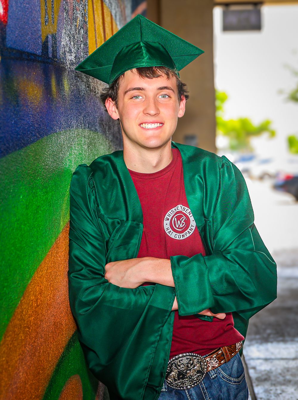 Graduate in emerald green cap and gown poses against colorful wall background.