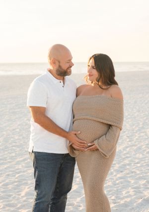 Expecting couple sharing tender moment on beach in matching white and beige outfits.