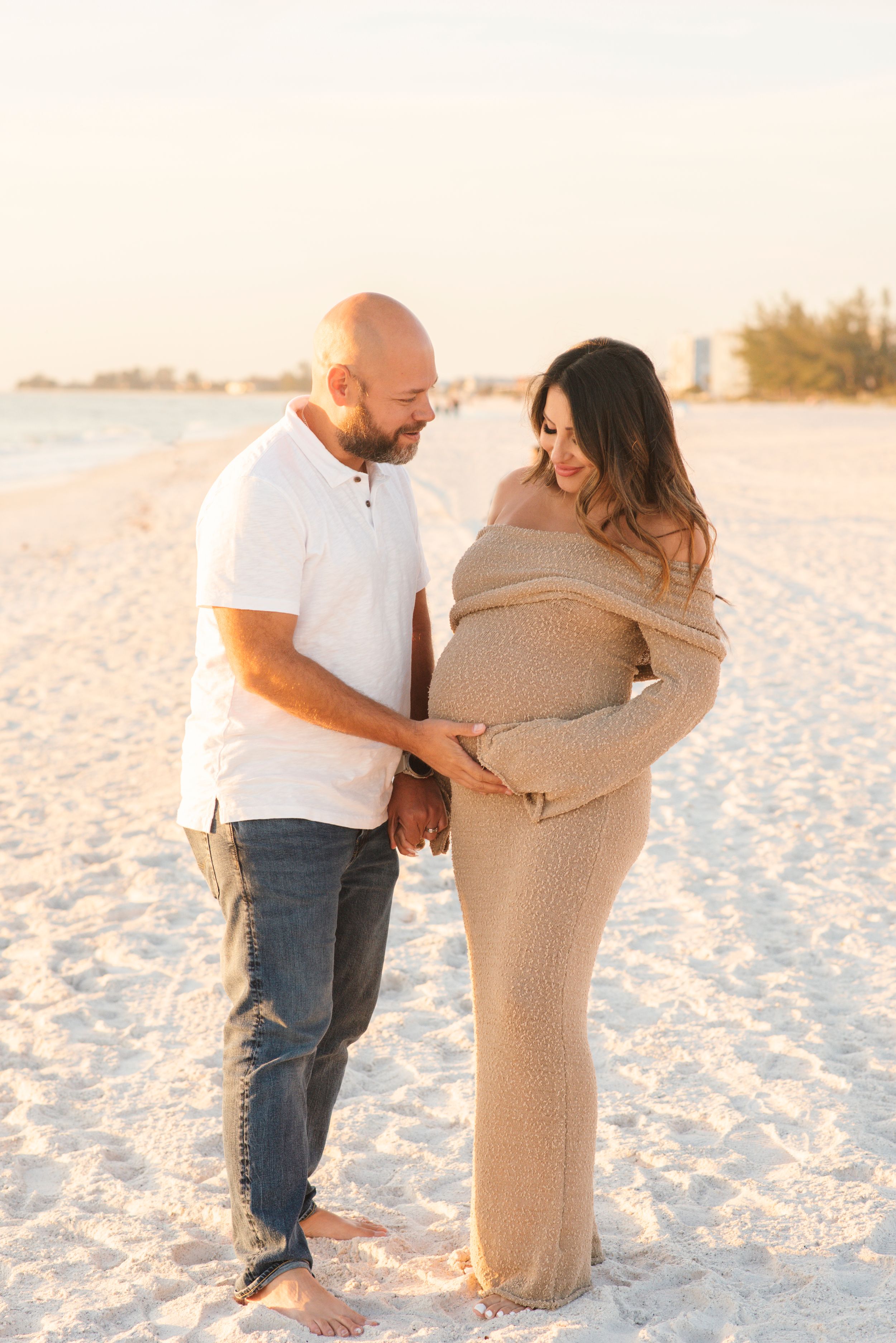 Maternity beach portrait of couple in casual attire standing on white sand.