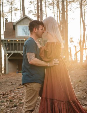 An intimate embrace between a couple in casual attire near a woodland treehouse at golden hour.