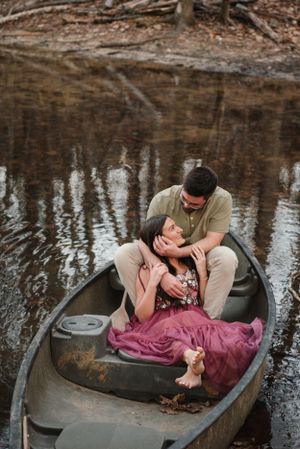 A romantic couple sits close together in a wooden canoe floating on calm water surrounded by nature.