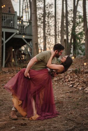 A romantic couple shares an intimate dip pose in front of a house at dusk, with her burgundy tulle skirt flowing.
