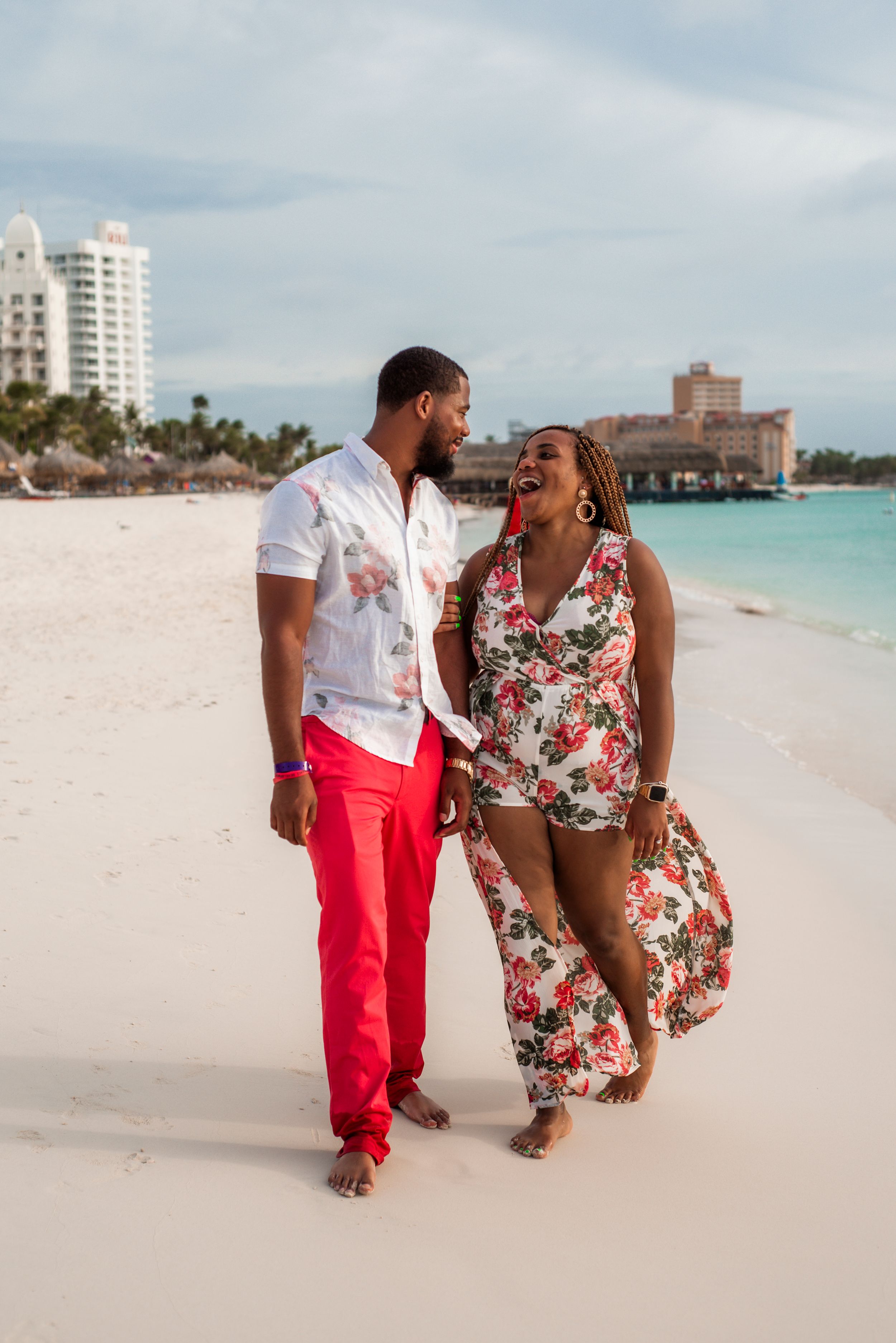Couple strolling barefoot on a tropical beach with high-rise buildings and palm trees in the background.