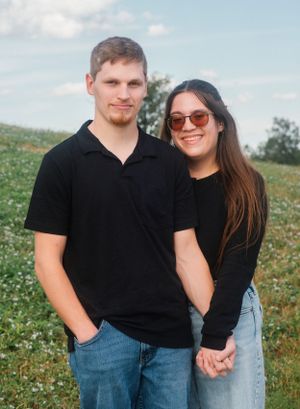 A casual couple poses together in matching black tops against a scenic hillside backdrop.