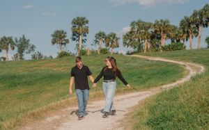 A couple walks hand in hand along a winding dirt path surrounded by green hills and palm trees.