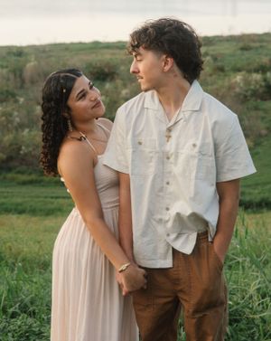 A couple embraces outdoors in coordinated outfits against a lush green hillside landscape.