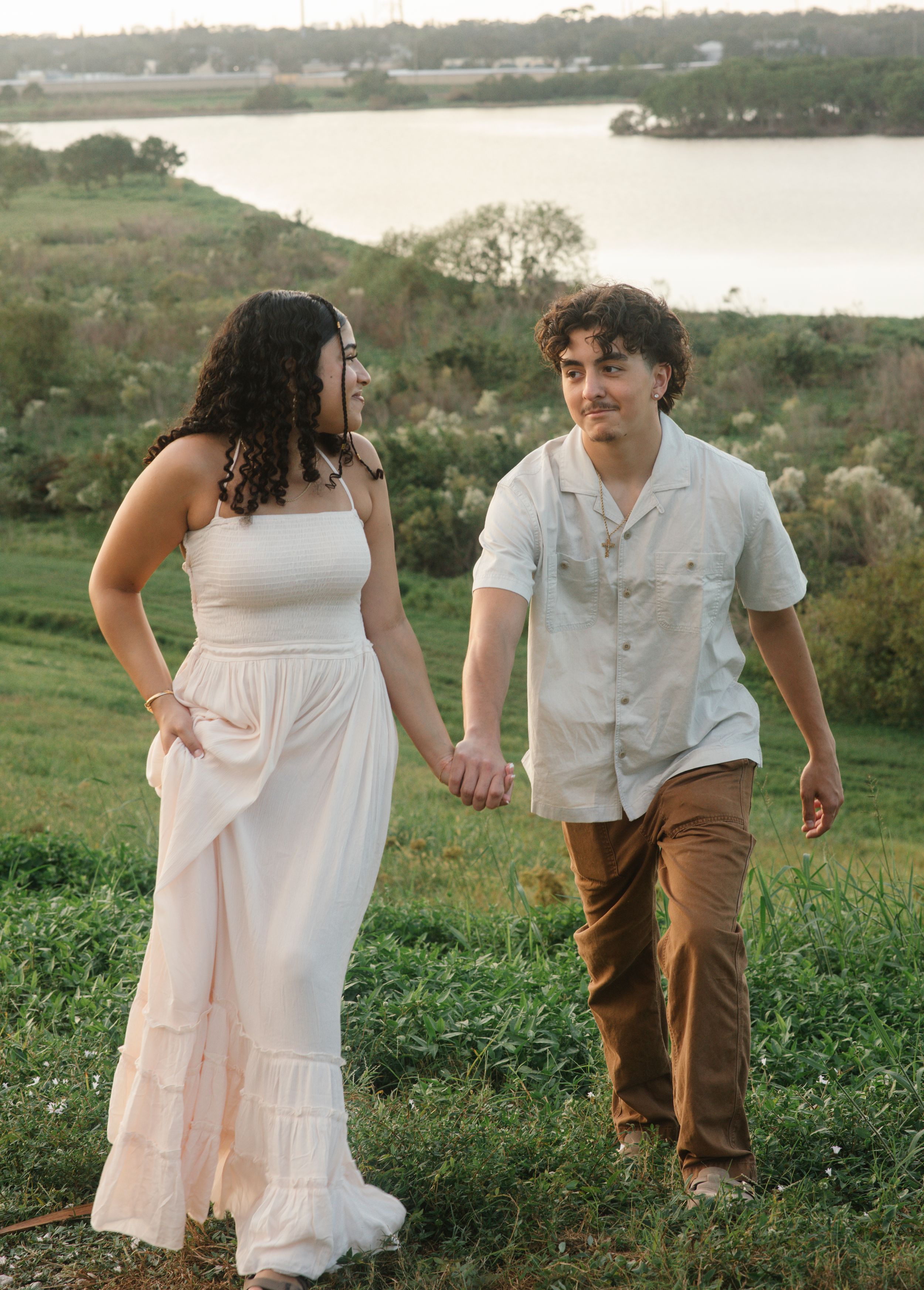 A couple shares a romantic moment on a grassy hillside overlooking a serene lake at sunset.