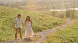 A couple walks along a winding dirt path through a sunlit grassy hillside overlooking a lake.