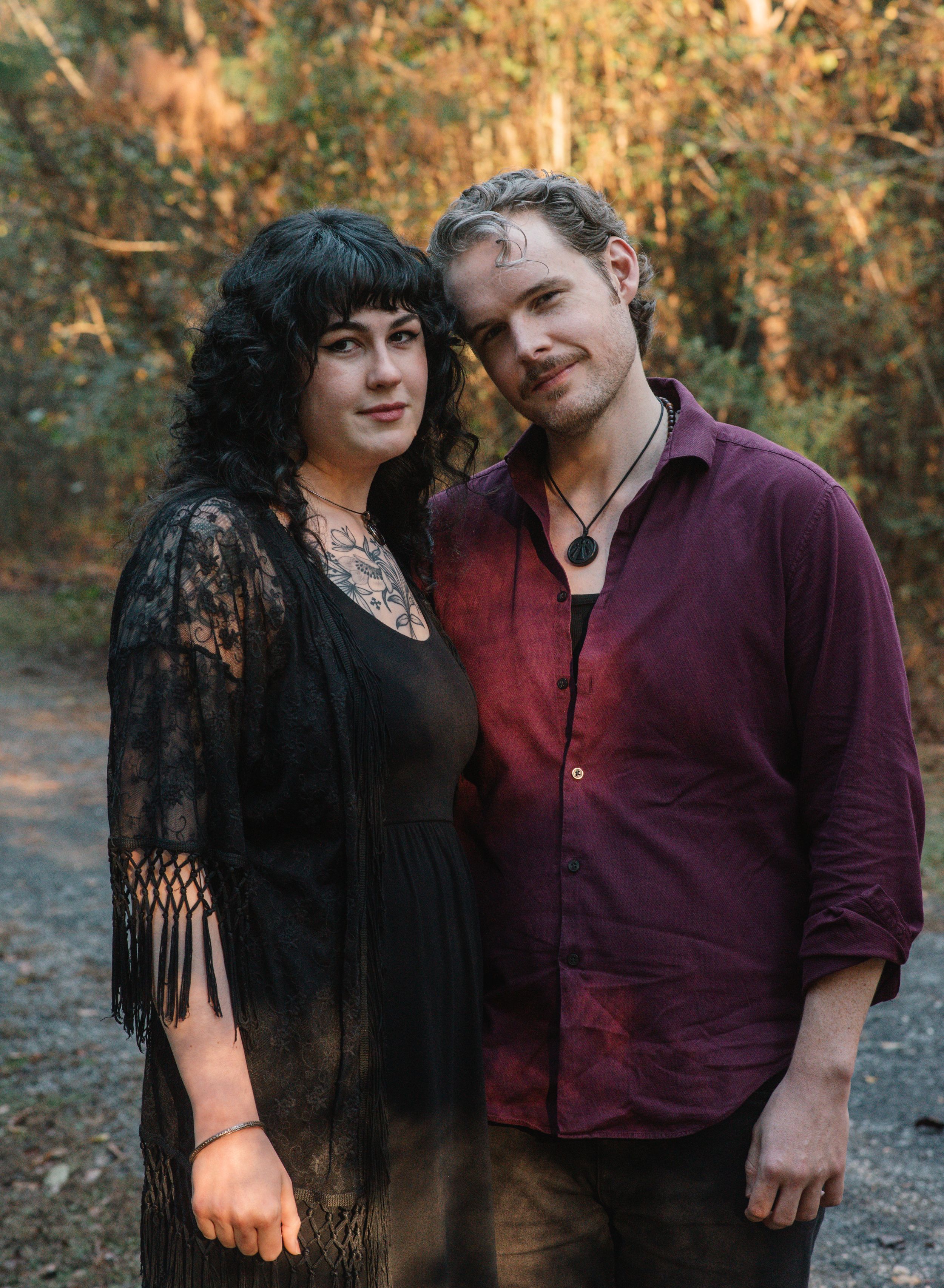 A couple in gothic-inspired outfits poses together for a portrait in an autumn forest setting.