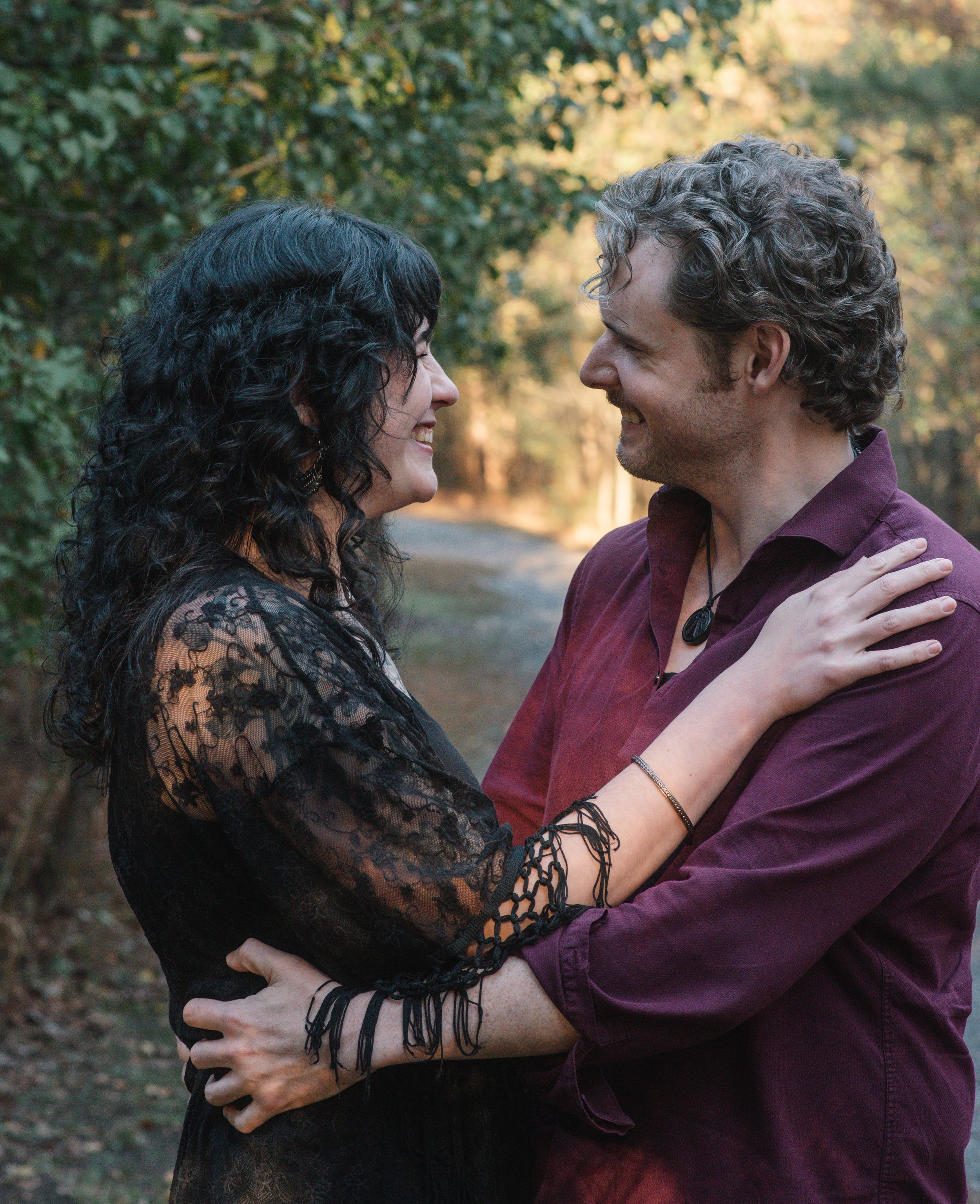Two people embrace while wearing dark romantic attire against an autumn forest backdrop.