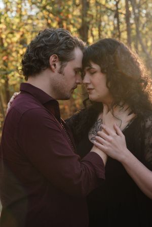 An intimate moment between two people in dark romantic attire with sunlit trees in the background.