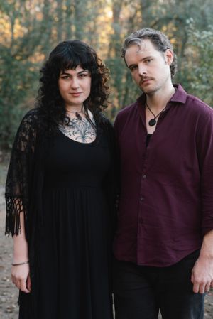 Portrait of a couple in coordinated dark outfits standing in a wooded setting.