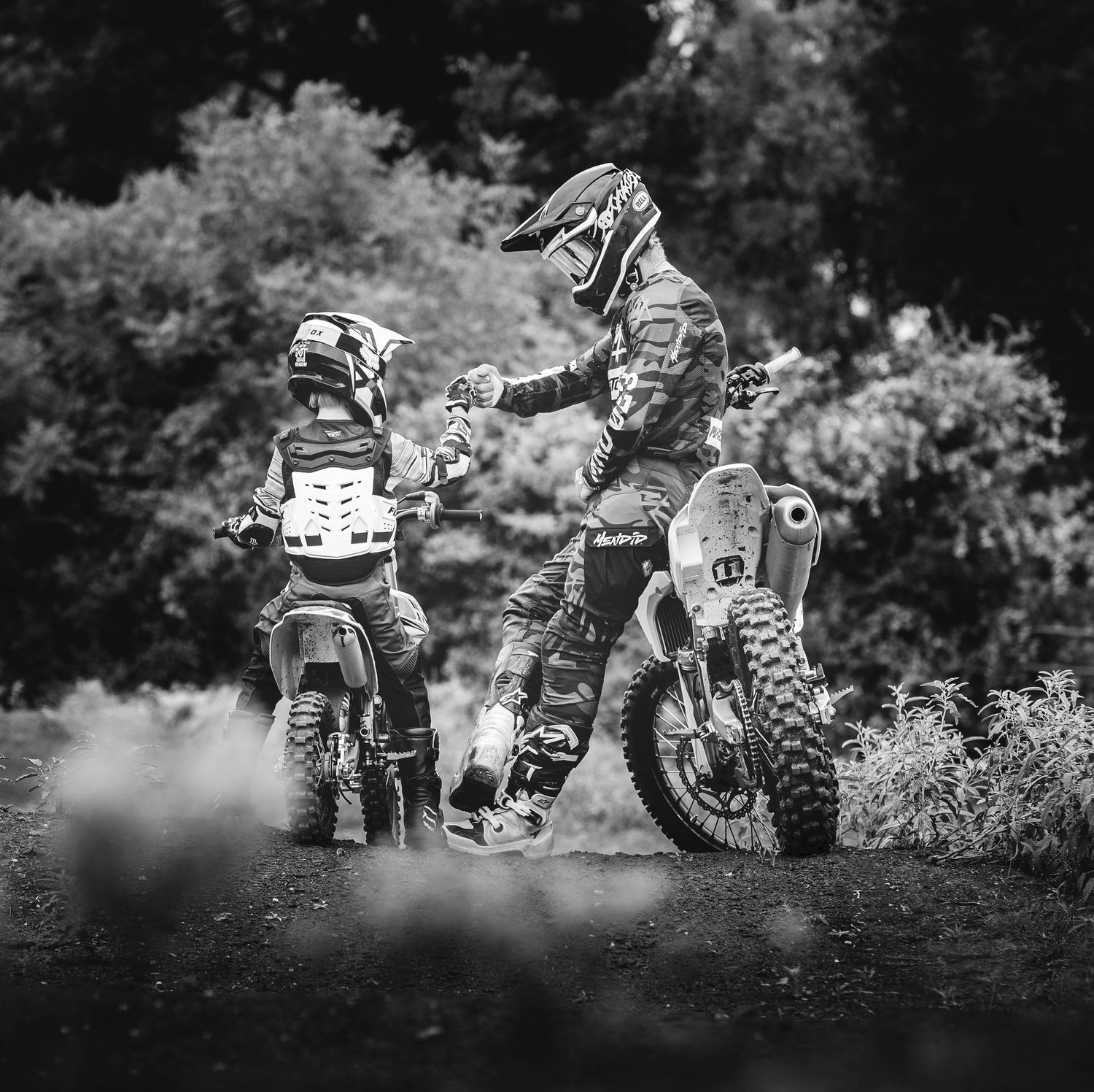 Two dirt bike riders fist bump on their motorcycles in a black and white forest scene.