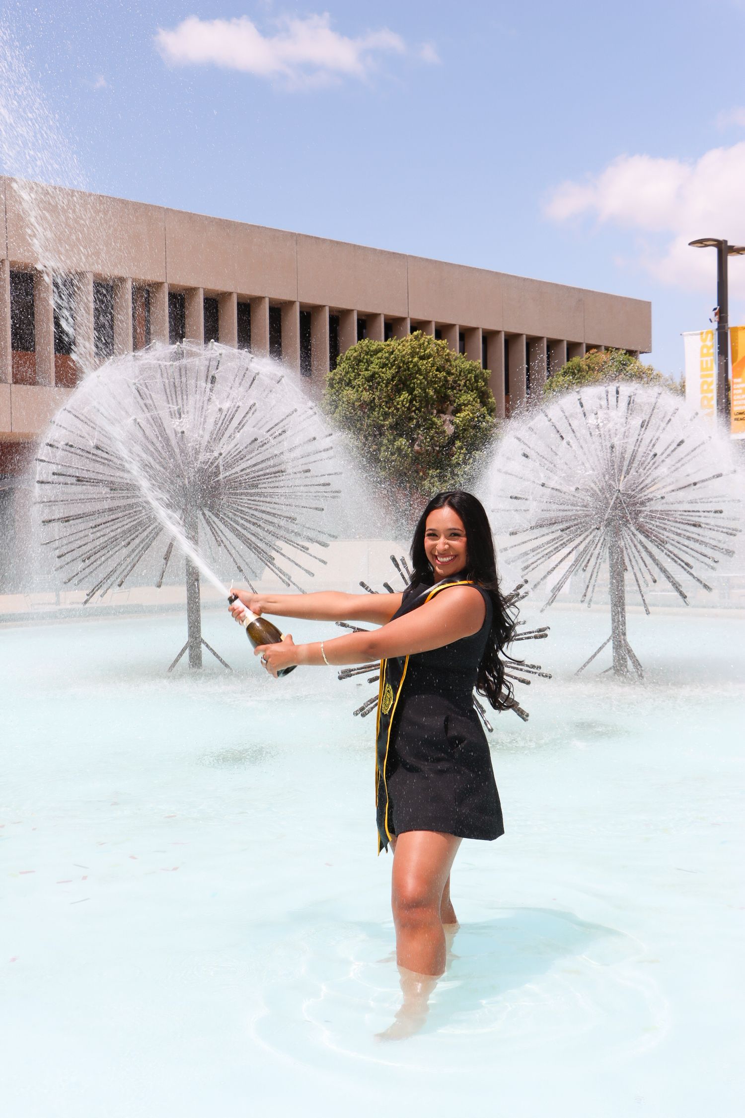 Graduate in black dress poses in fountain with water features spraying in background.
