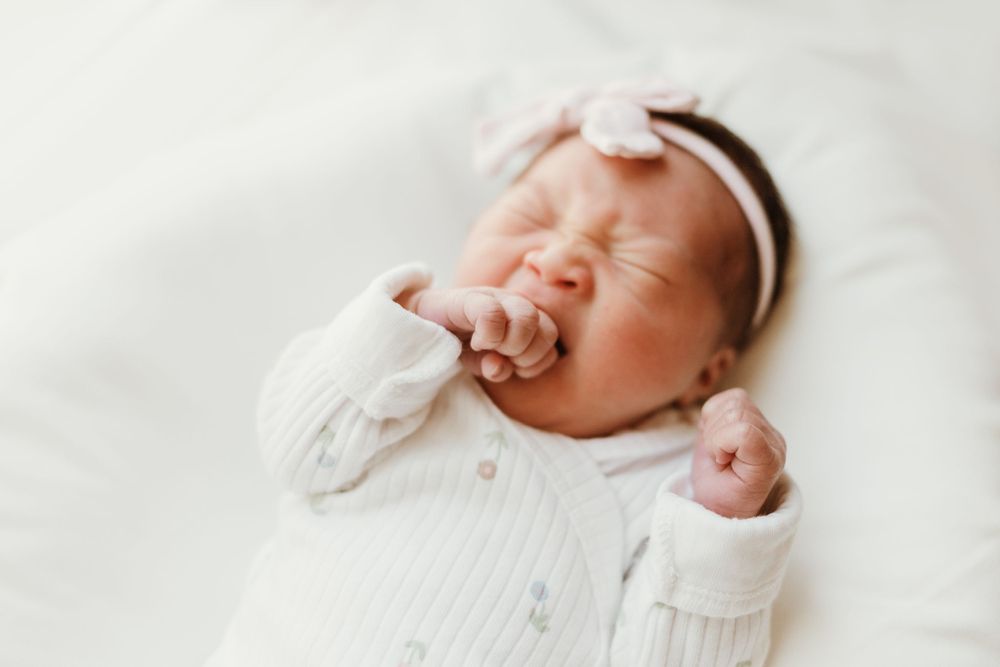 Newborn baby yawns while wearing white outfit and headband on white bedding.