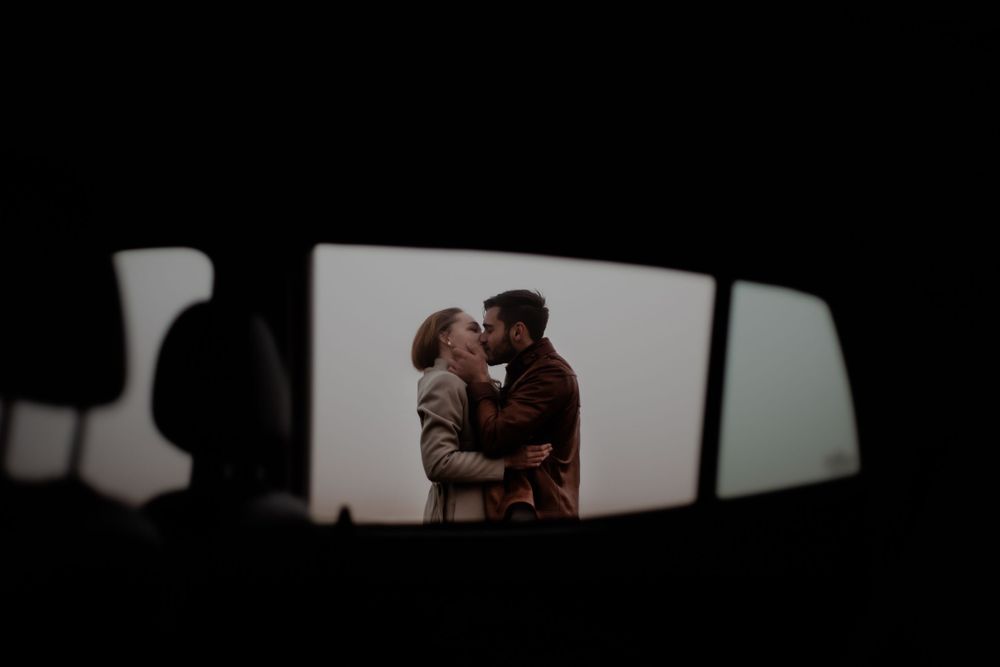 A silhouetted couple's tender moment viewed through a car's rearview mirror at sunset.