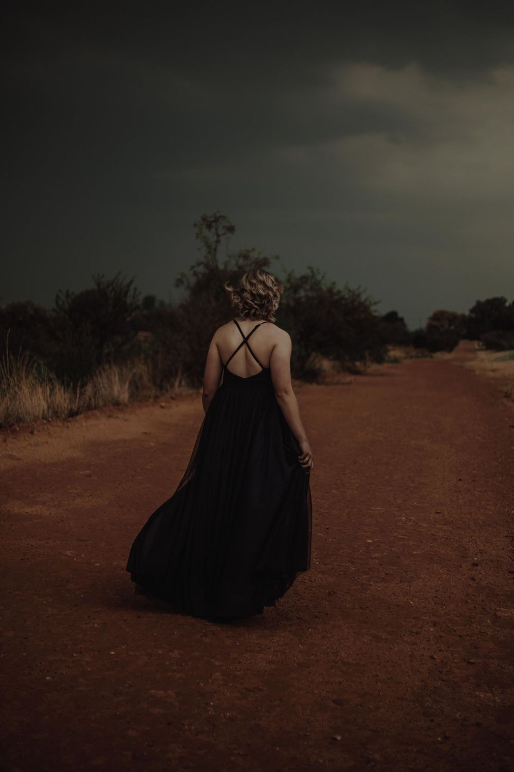 A figure in a flowing black dress walks down a rustic dirt path under stormy skies.